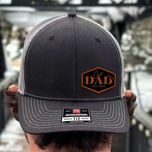 May include: A black trucker hat with a gray mesh back and a white stitched brim. The hat features a brown patch with the words "Girl Dad EST. 2025". The hat is adjustable and has a Richardson 112 Trucker label.