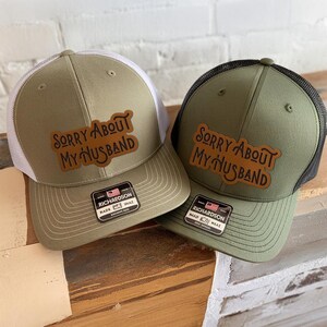 May include: Two olive green trucker hats with the text "SORRY ABOUT MY HUSBAND" on a brown leather patch. One hat has a white mesh back, the other has a black mesh back. Both hats have a Richardson tag.