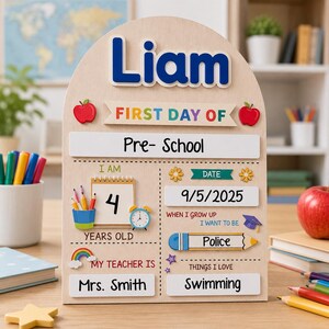 Puede incluir: Un letrero de madera arqueado para el primer día de clases con el nombre Liam en letras azules. Incluye secciones para grado, edad, fecha, maestro, meta profesional e intereses, decorado con iconos coloridos como manzanas, lápices y un arcoíris.