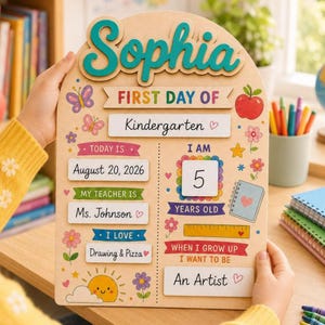 Puede incluir: Un letrero de madera en forma de arco para el primer día de clases con el nombre Sophia en color verde azulado. El tablero incluye secciones para fecha, maestro, edad e intereses con ilustraciones coloridas de mariposa, manzana y sol.