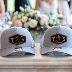 May include: Two gray trucker hats with a black and gold patch featuring the letters "T & S" and the date "10.19.2024". The hats have a mesh back and a curved brim.