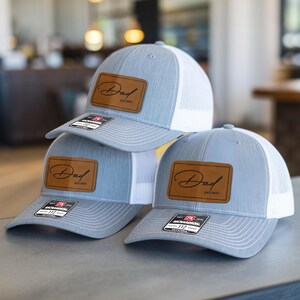 May include: Three gray and white trucker hats with a brown leather patch that says "Dad est. 2014".