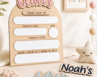 May include: An arched wooden sign with the name Olivia's in pink script at the top. It features blank white spaces for First Day Of, Today's Date, Age, Things I Love, and When I Grow Up, I Want To Be A. Two additional name plaques read Noah's in navy and Lily's in light blue.
