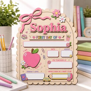 Puede incluir: Un letrero de madera clara con bordes festoneados, que presenta el nombre Sophia en letras rosas, una manzana rosa grande y secciones para detalles escolares como fecha, altura, profesor e intereses, decorado con lazos y flores rosas.
