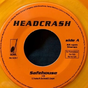 May include: Orange vinyl record with the text "HEADCRASH" in black. The label also includes the text "Safehouse" with the song length "3:58" and the songwriters "U. Franke/N. Berthold/S. Cooper". The record is a promotional copy, indicated by the text "For Promotional Use Only Not for Sale".