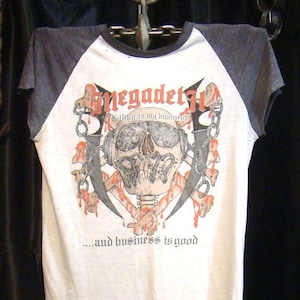 Puede incluir: Una camiseta blanca con mangas raglán negras y grises. La parte delantera de la camiseta presenta una calavera con huesos cruzados y el texto "Megadeth" en rojo y negro. El texto "Killing is my business... and business is good" está impreso debajo de la calavera.