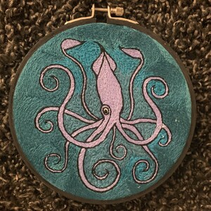 May include: Embroidered artwork featuring a lavender squid design on a teal background. The squid has detailed tentacles and a central body, all outlined in black. The embroidery is set in a black hoop, creating a finished piece of textile art.