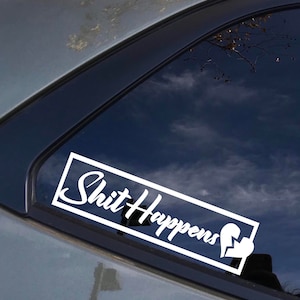 May include: White vinyl sticker with the text "Shit Happens" in a cursive font with a broken heart symbol at the end. The sticker is on the back window of a silver car.