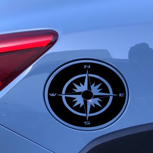 May include: A black compass sticker with a white compass rose and the letters N, S, E, and W on a blue car.