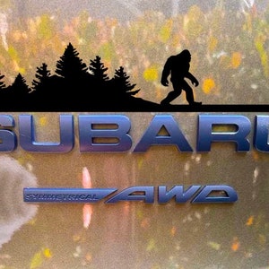 May include: A black silhouette of a Bigfoot walking past a line of trees, with the word "SUBARU" in blue letters below. The words "SYMMETRICAL AWD" are also visible in blue letters below the word "SUBARU".
