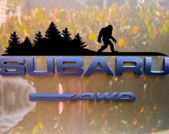 May include: A black silhouette of a Bigfoot walking past a line of trees, with the word "SUBARU" in blue letters below. The words "SYMMETRICAL AWD" are also visible in blue letters below the word "SUBARU".