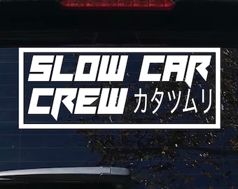 May include: White sticker with black outline on a black car window. The sticker says "SLOW CAR CREW カツリ".