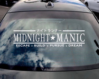 Könnte beinhalten: Ein weißer Vinyl-Aufkleber mit dem Text "MIDNIGHT★MANIC" und den Worten "ESCAPE • BUILD • PURSUE • DREAM" darunter. Der Aufkleber befindet sich auf der Heckscheibe eines schwarzen Autos.