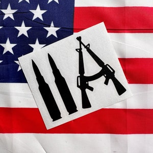 May include: A white sticker with a black silhouette of two rifles and three bullets. The sticker is on top of an American flag.