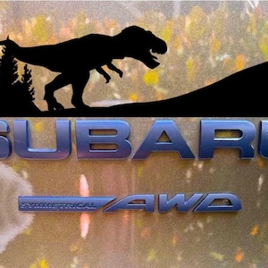 Puede incluir: Logotipo de Subaru azul met&aacute;lico con el texto &laquo;SUBARU&raquo; y &laquo;SYMMETRICAL AWD&raquo;. Encima del logotipo, una silueta negra de un Tyrannosaurus Rex y un paisaje monta&ntilde;oso con pinos.