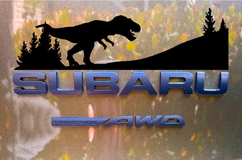 Puede incluir: Logotipo de Subaru azul met&aacute;lico con el texto &laquo;SUBARU&raquo; y &laquo;SYMMETRICAL AWD&raquo;. Encima del logotipo, una silueta negra de un Tyrannosaurus Rex y un paisaje monta&ntilde;oso con pinos.