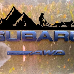 May include: A black silhouette of a mountain range with a person and dogs pulling a sled. The text "SUBARU" is in blue letters below the silhouette. The text "SYMMETRICAL AWD" is in blue letters below the text "SUBARU".