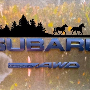 May include: A metallic blue Subaru emblem with the text "SUBARU" and "SYMMETRICAL AWD". Above the text is a black silhouette of a forest and two horses. The background has a blurred, natural color.