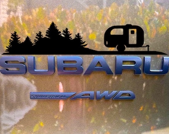 May include: A black Subaru emblem with the word "SUBARU" in large letters. Below the word is a smaller emblem that says "SYMMETRICAL AWD". Above the word "SUBARU" is a silhouette of a forest with a small camper trailer.