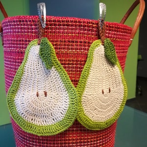 Könnte beinhalten: Ein roter gewebter Korb mit zwei gehäkelten birnenförmigen Topflappen, die oben befestigt sind. Die Topflappen sind grün und weiß mit einem lächelnden Gesicht.