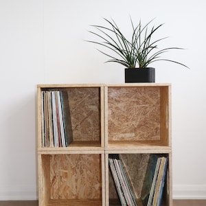 May include: A four-cube storage unit made from particle board, with a plant in a black pot on top. Two cubes contain vinyl records. The unit is light brown and sits against a white wall. The unit is approximately 76cm high.