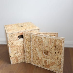 Könnte beinhalten: Eine Holzkiste mit Deckel und Griff sowie drei Holzplatten. Die Kiste und die Platten bestehen aus hellbraunen OSB-Platten mit sichtbaren Holzspänen. Jede Platte hat ein kreisförmiges Loch.