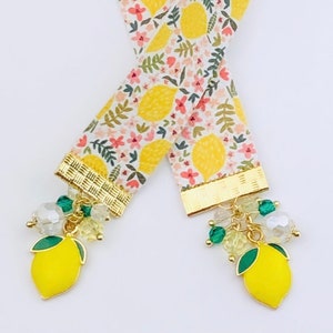 May include: A white ribbon bookmark with a lemon and floral pattern. The bookmark has gold trim and a lemon charm with green leaves and clear and green beads.