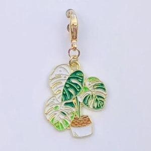 May include: A gold-colored charm featuring a green and white monstera plant in a white pot. The charm is attached to a gold clasp. The text "Duchess Gems STUDIO" is printed at the top of the white card.