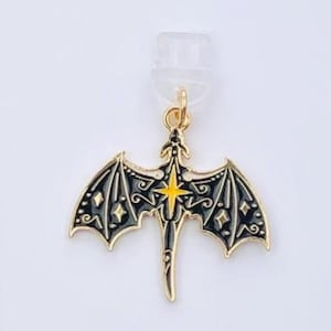 May include: A gold-colored dragon pendant with black enamel detailing and a yellow starburst design. The pendant is attached to a white card with the text "Duchess Gems STUDIO".