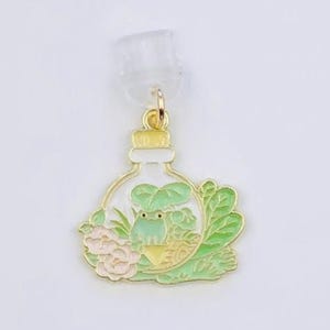 May include: A gold-colored terrarium charm with a frog and plants inside a glass bottle. The charm is attached to a clear earring back. The text "Duchess Gems STUDIO" is printed on the white card.