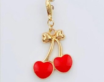 May include: A gold-coloured charm featuring two red cherries with a gold bow. The charm is attached to a gold clasp and is displayed on a white card with the text "Duchess Gems STUDIO".