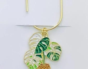 May include: A gold-colored metal bookmark with a monstera plant charm. The charm features green and white leaves in a white pot. The bookmark is attached to a white card with the text "Duchess Gems".