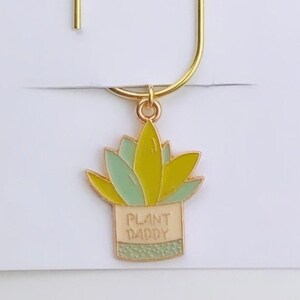 May include: A gold-colored bookmark with a plant charm. The charm is a yellow and green succulent in a tan pot, with the words "PLANT DADDY" on the pot. The bookmark is on a white card with the text "Duchess Gems STUDIO".