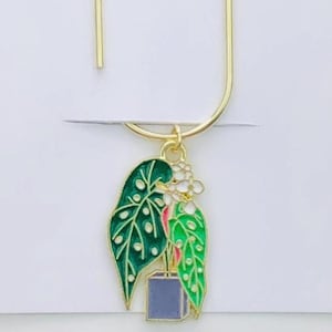 May include: A gold-colored metal bookmark with a plant charm. The charm features green and red leaves with white accents and a blue square pot. The bookmark is attached to a white card with the text "Duchess Gems STUDIO".