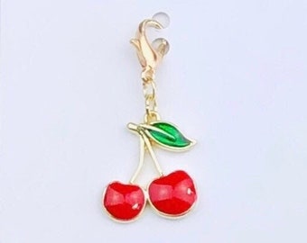 May include: Gold charm featuring a red cherry design. The charm has a green leaf and a gold clasp. Duchess Gems Studio.
