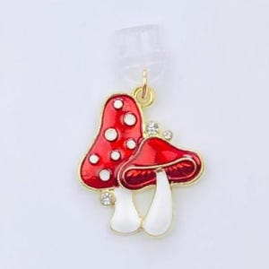 May include: A pair of red mushroom earrings with white spots and white stems. The earrings have a gold-colored metal finish and are embellished with small, clear crystals. The earrings are displayed on a white card with the text "Duchess Gems STUDIO".