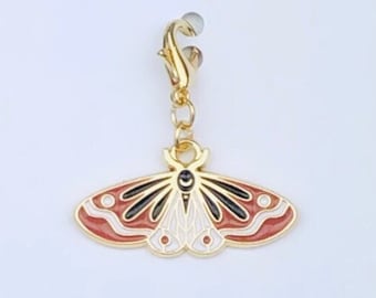 May include: Gold-tone charm featuring a red, white, and black moth design. The charm has a lobster clasp for attaching to a bag, keychain, or other accessories. The charm is displayed on a white card with the text "Duchess Gems Studio".