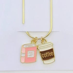 May include: A gold-coloured metal bookmark featuring a pink book charm and a coffee cup charm. The coffee cup is brown and white with the word "coffee". The bookmark is on a white card with the text "Duchess Gems STUDIO".