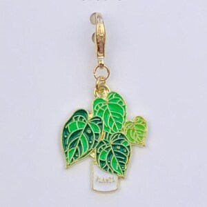 May include: A gold-colored charm featuring a green plant in a white pot. The plant has detailed green leaves with dark green outlines. The charm is attached to a gold-colored clasp. The text "Duchess Gems STUDIO" is at the top.