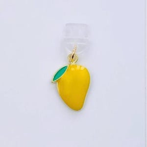 May include: A yellow mango charm with a green leaf, hanging from a gold-colored bail. The charm is on a white card with the text "Duchess Gems STUDIO". The mango is a bright yellow color.