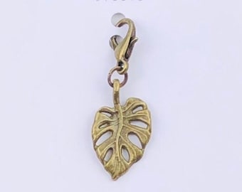 May include: Gold-toned metal leaf charm with a lobster clasp. The charm is shaped like a monstera leaf and is attached to a white card with the text "Duchess Gems Studio".