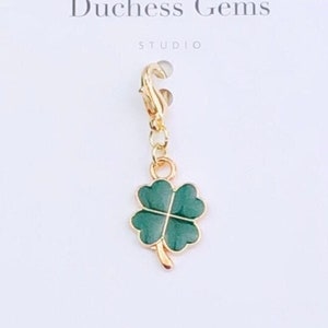 May include: A gold charm featuring a green four-leaf clover design. The charm is attached to a gold chain with a lobster clasp. The charm is displayed on a white background with the text "Duchess Gems Studio".