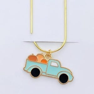 Peut inclure: Marque-page en métal doré avec un charm en forme de camion bleu clair transportant des citrouilles oranges. Le marque-page est fixé à une carte blanche avec le texte "Duchess Gems STUDIO". Le charm du camion a des roues noires et un contour noir.