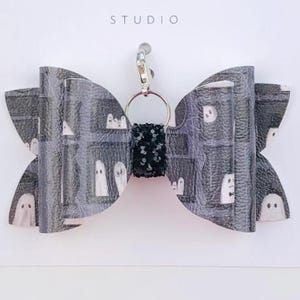 May include: A Halloween-themed bow-shaped keyring with a gray and white ghost pattern. The bow is attached to a silver-coloured metal clasp and a black and white patterned centre. The text "Duchess Gems STUDIO" is printed on the white card.