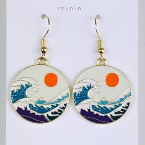 May include: Gold-tone earrings with a circular design. Each earring features a white background, an orange sun, and a wave pattern in blue and purple. The earrings are presented on a white card with the text "Duchess Gems STUDIO".