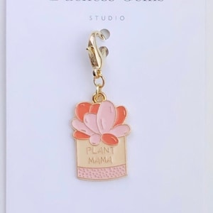 May include: A gold charm with a pink and orange plant in a pot. The pot has the words "Plant Mama" on it. The charm is attached to a gold clip.
