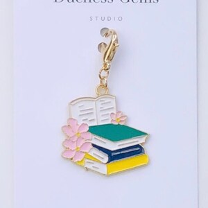May include: Gold charm featuring a stack of books with a green, blue, and yellow cover. The top book is open with white pages and pink flowers. The charm has a gold clasp.
