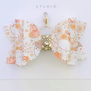Könnte beinhalten: Eine weiße Schleife mit einem goldglitzernden Mittelteil und einem goldenen Verschluss. Die Schleife hat ein sich wiederholendes Muster aus Kürbissen, Blumen und Spinnen. Der Text "Duchess Gems STUDIO" ist auf die weiße Karte gedruckt.