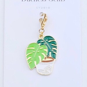 May include: A gold-colored charm featuring a green and white Monstera plant in a white pot. The charm has a lobster clasp and the text "Duchess Gems Studio" is printed on the card.
