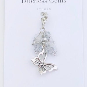 May include: A silver charm with a butterfly design and clear crystal beads. The charm is attached to a silver loop. The charm is labeled "Duchess Gems Studio".